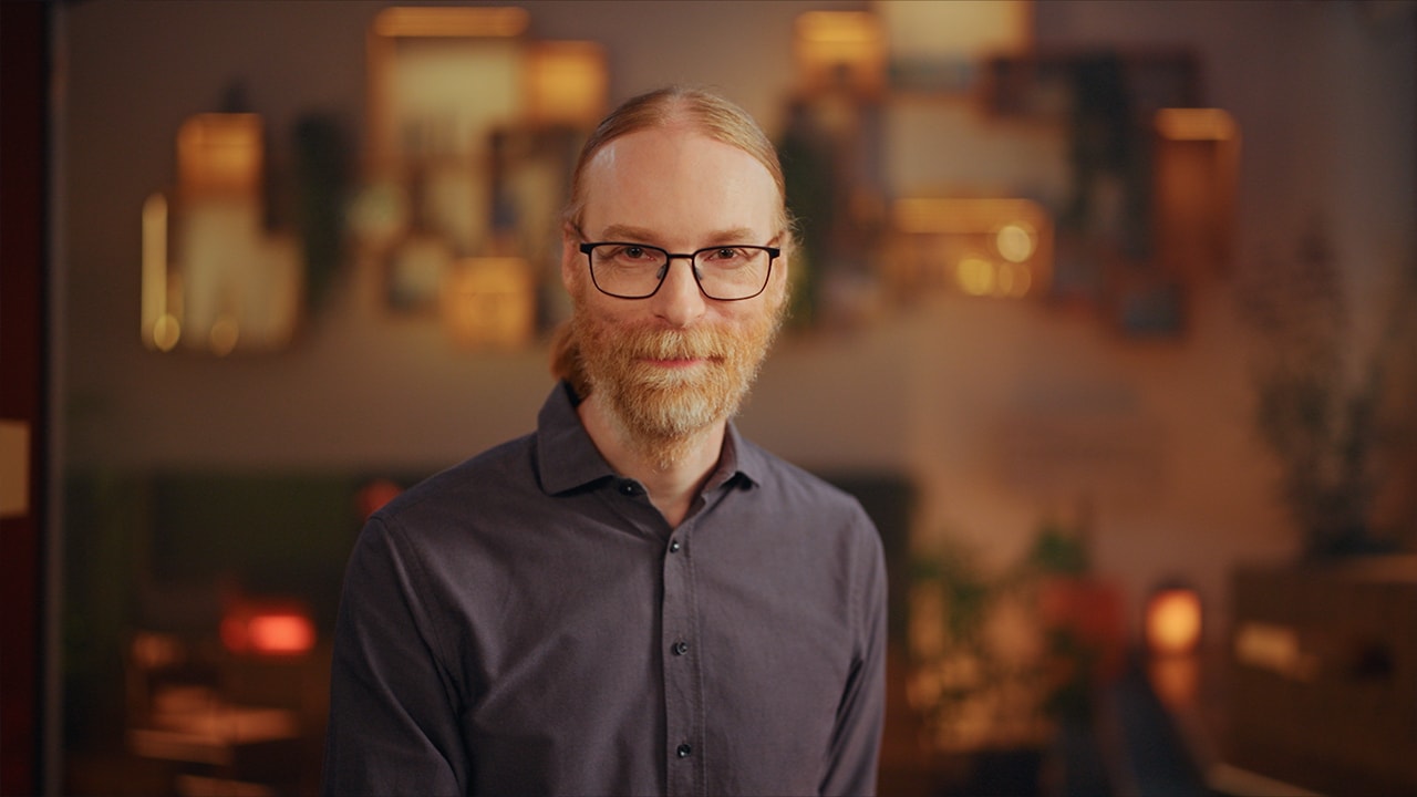 Portrait of a bearded man, Jens Bergenström, wearing glasses, standing against a softly lit background.