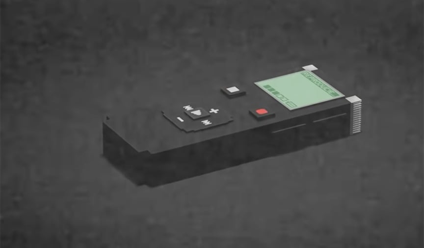 A blocky black digital voice recorder with a green screen and a red record button lies on a dark grey surface.